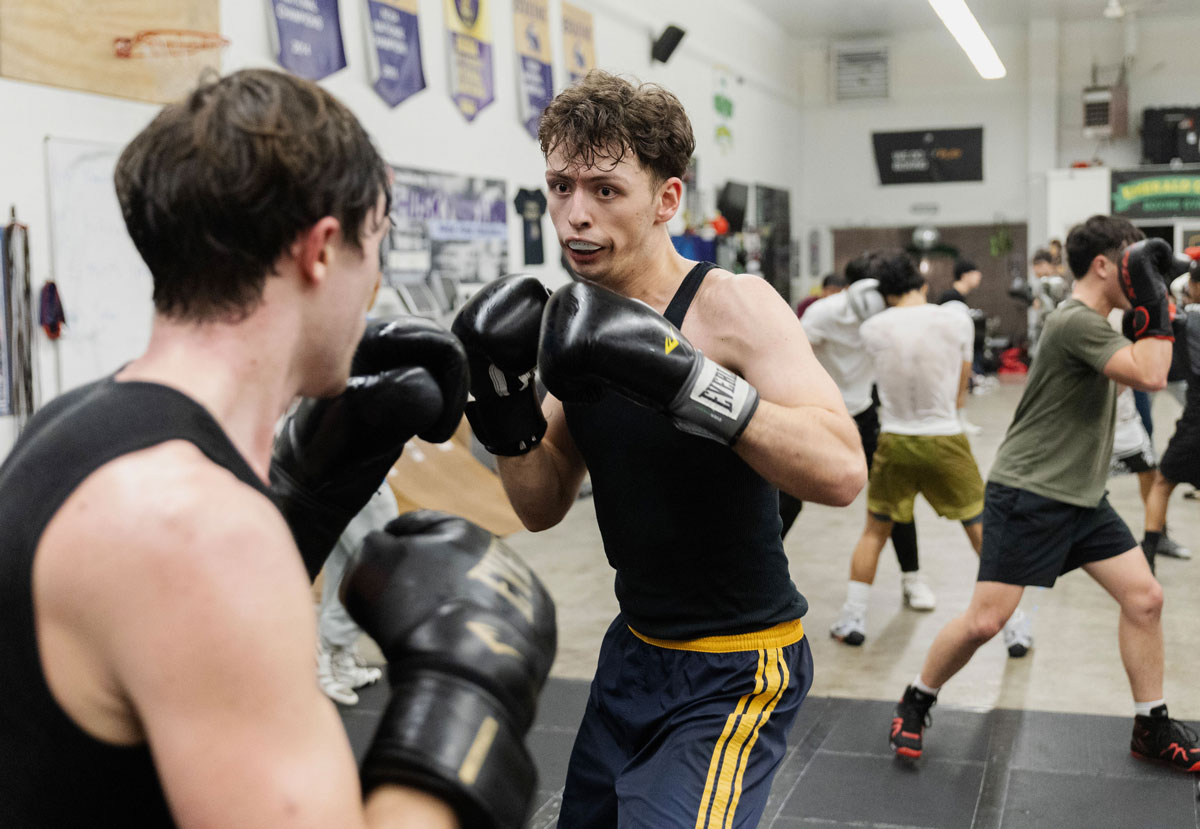 A young man wearing a mouthguard spars with another young man in a boxing gym.