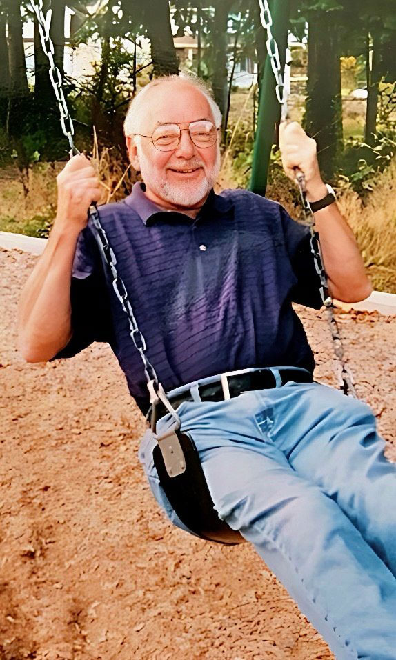 A man in a purple polo shirt and jeans swings on a swingset at a playground.