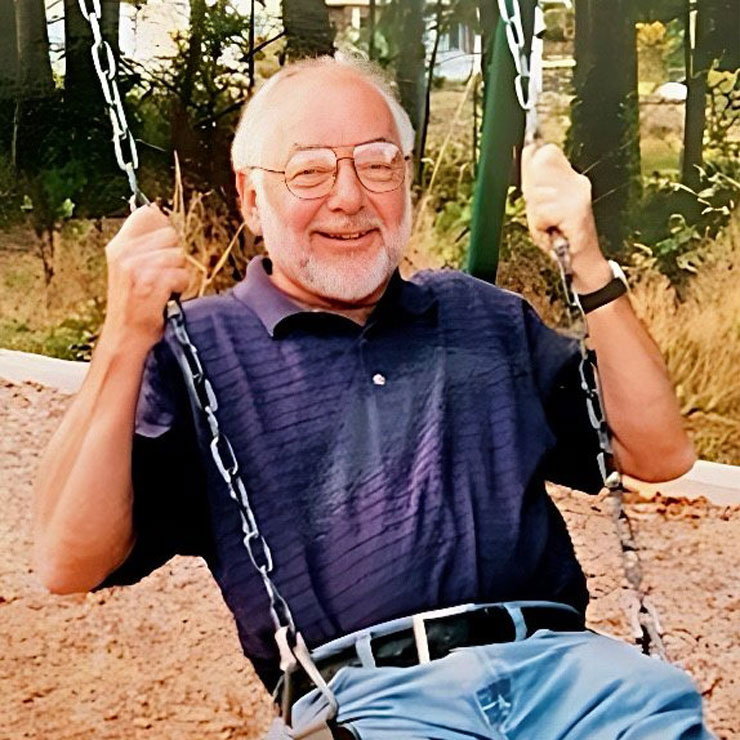 A man in a purple polo shirt and jeans swings on a swingset at a playground.