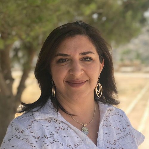 A woman with olive skin and brown hair in a white blouse stands on a dusty road smiling.