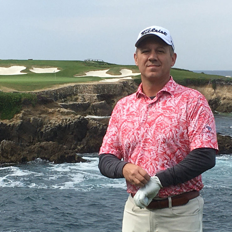 A man in a pink golf shirt stands on the shore of an island with a golf course in the background.