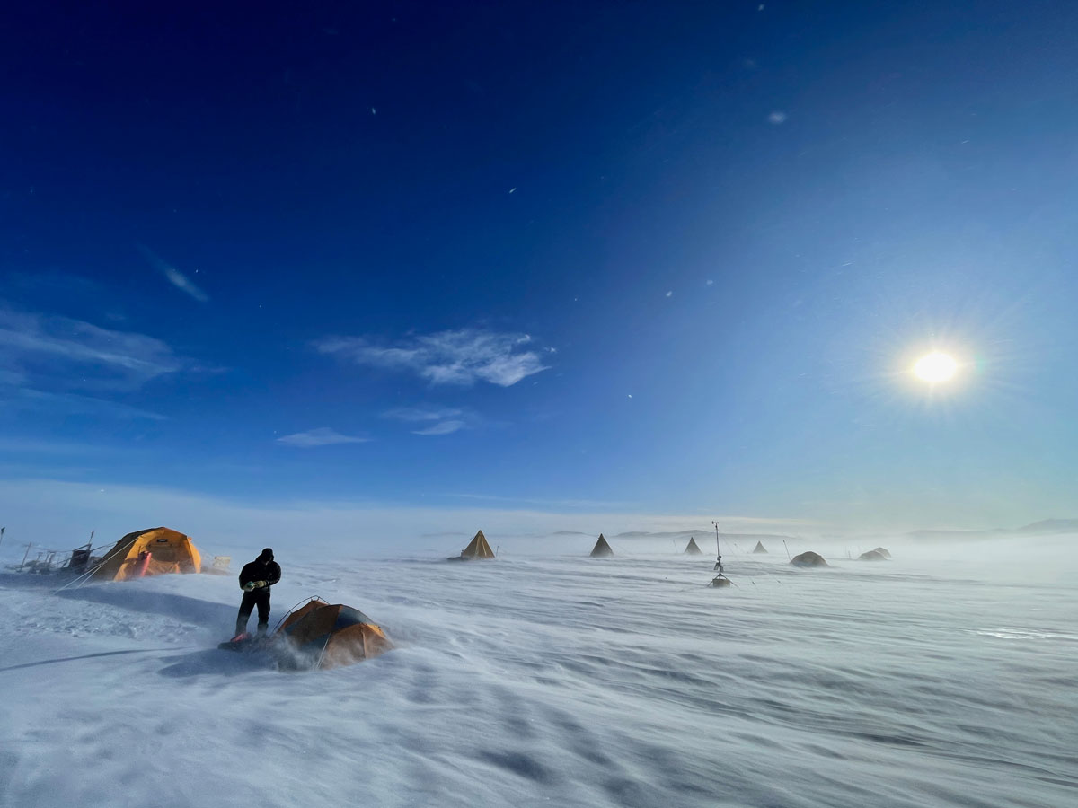 A windswept snowfield with a few people near small tents against a bright blue sky in Antarctica