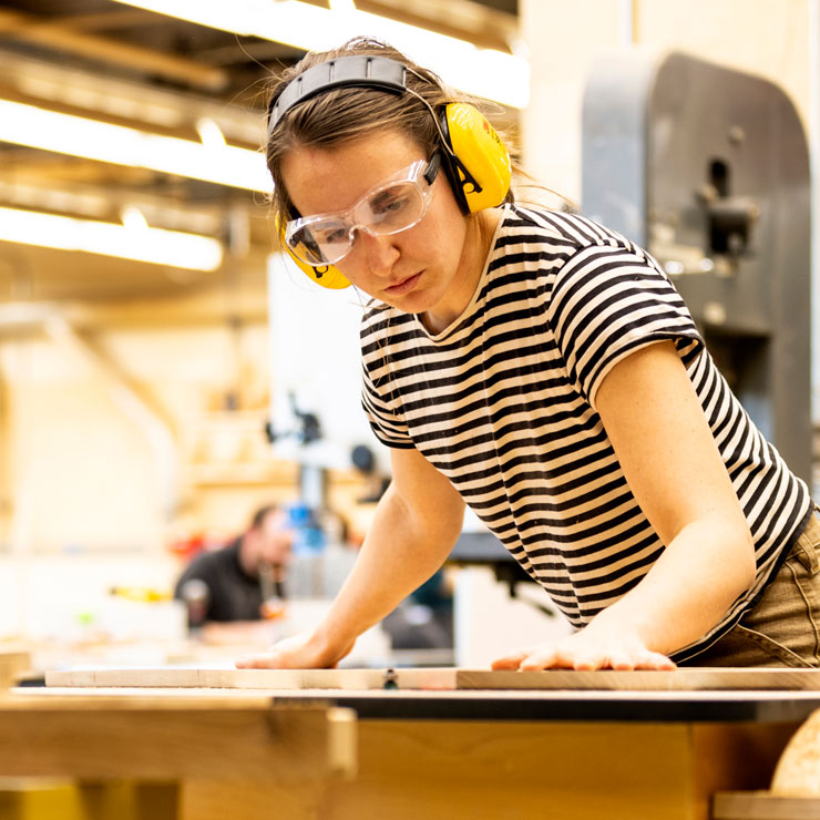 A woman in a striped shirt and protective gear steadies a piece of wood in a workshop