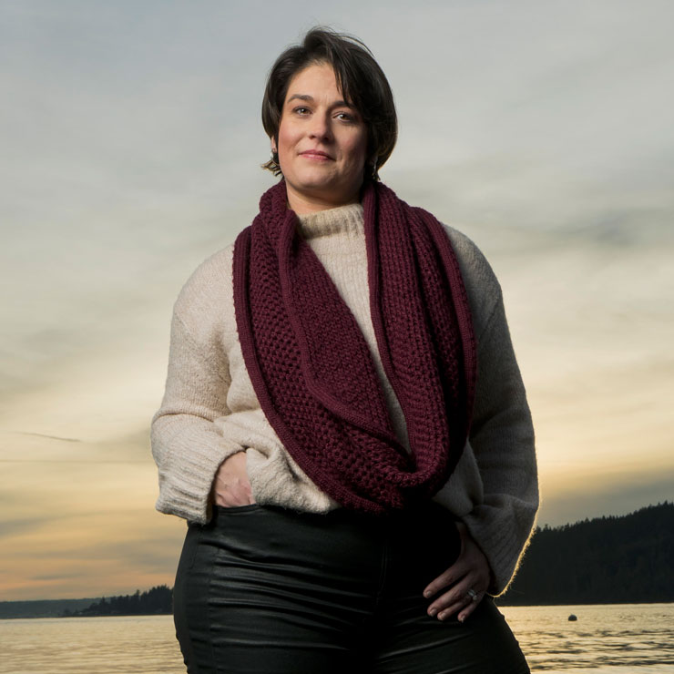 A woman in a beige sweater and red scarf stands confidently at the seaside