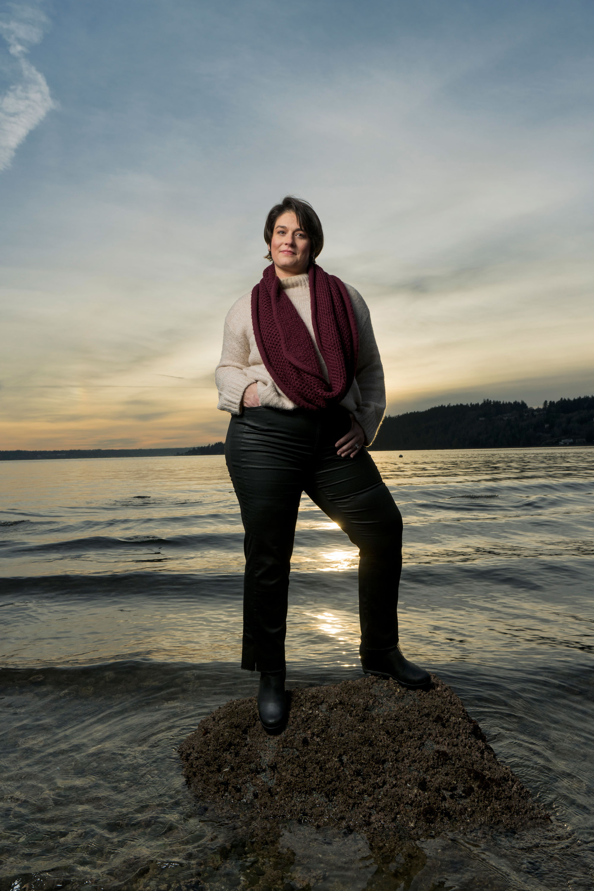 A woman in a beige sweater and red scarf stands confidently on a rock at the seaside