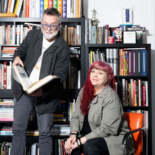 A man and woman smile in front of a bookshelf in their home