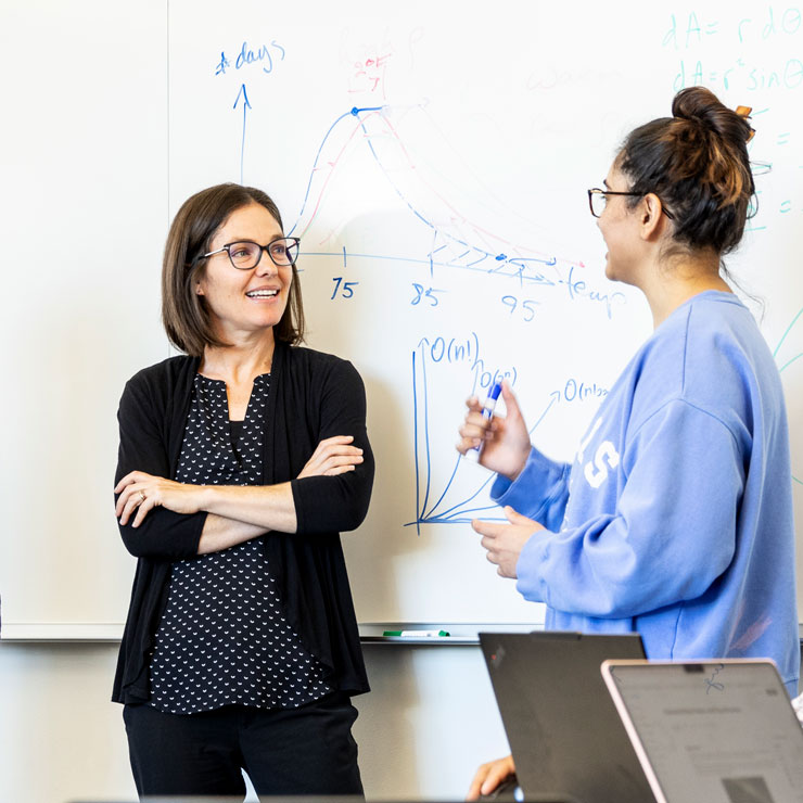 A woman in a polka dot blouse and black cardigan folds her arms while chatting with a student in a baggy blue sweatshirt.