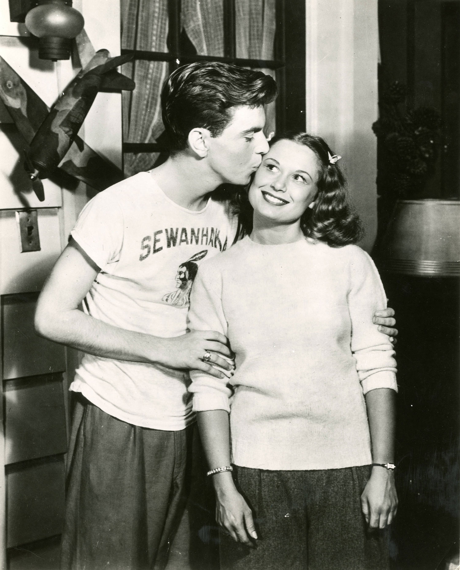 A 1930s photo shows a woman in a sweater smiling and being kissed on the cheek by a man.