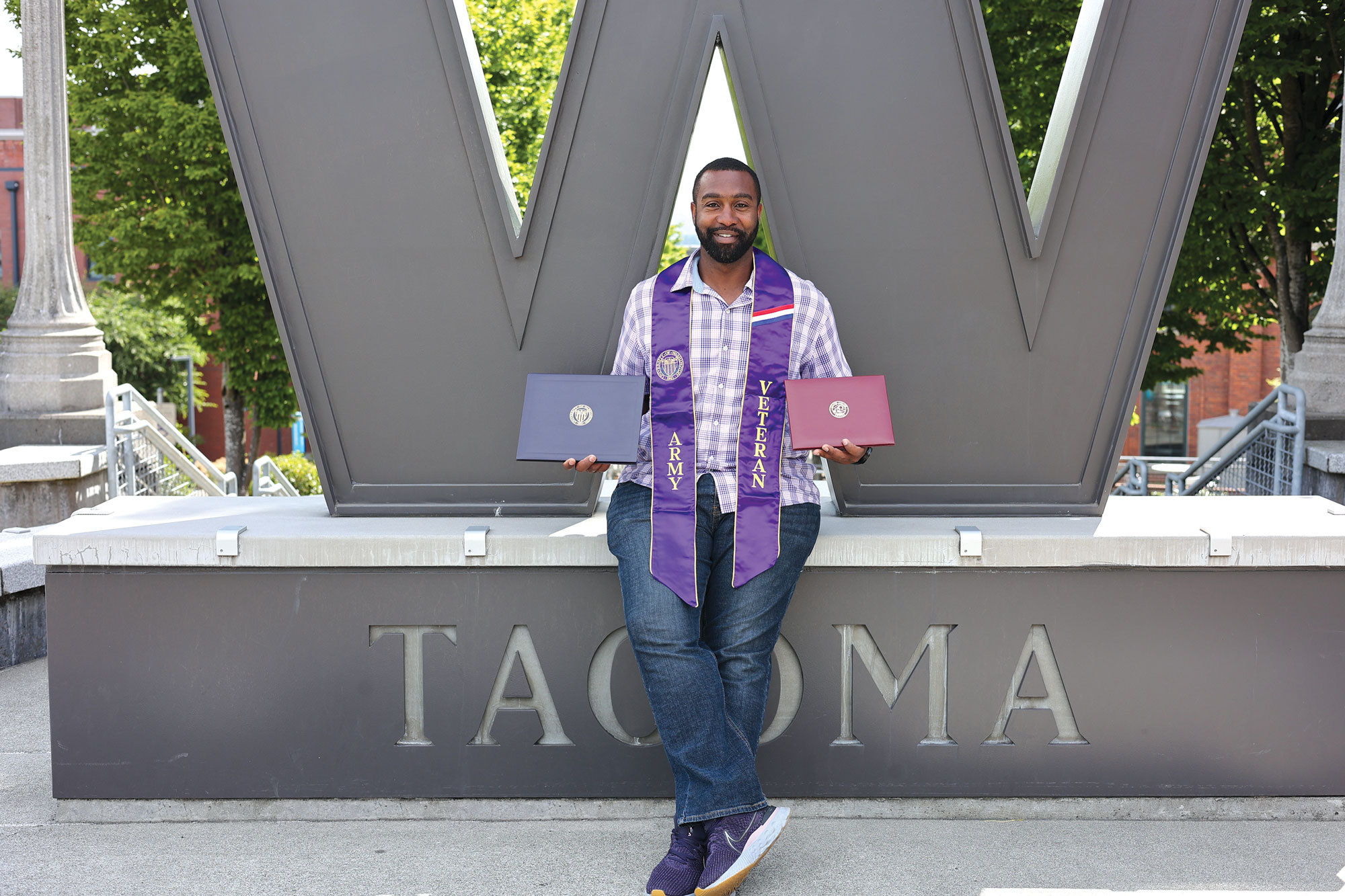 A man wearing an academic stole that reads "Army Veteran" holds his degrees in front of a large W statue.