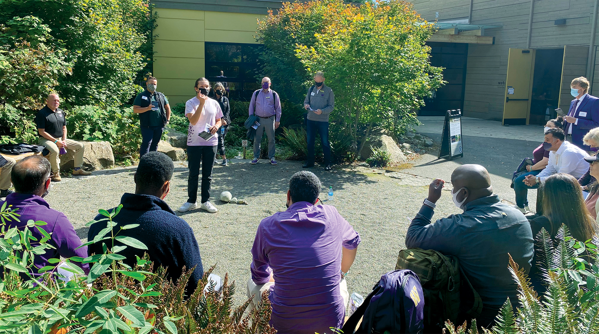 A young man gives a speech to a group of formally dressed people in an outdoor plaza