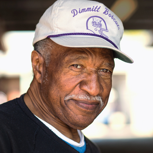 Junior Coffey smiles, wearing a black shirt and a white hat that says "Dimmitt Bobcats" in purple lettering.