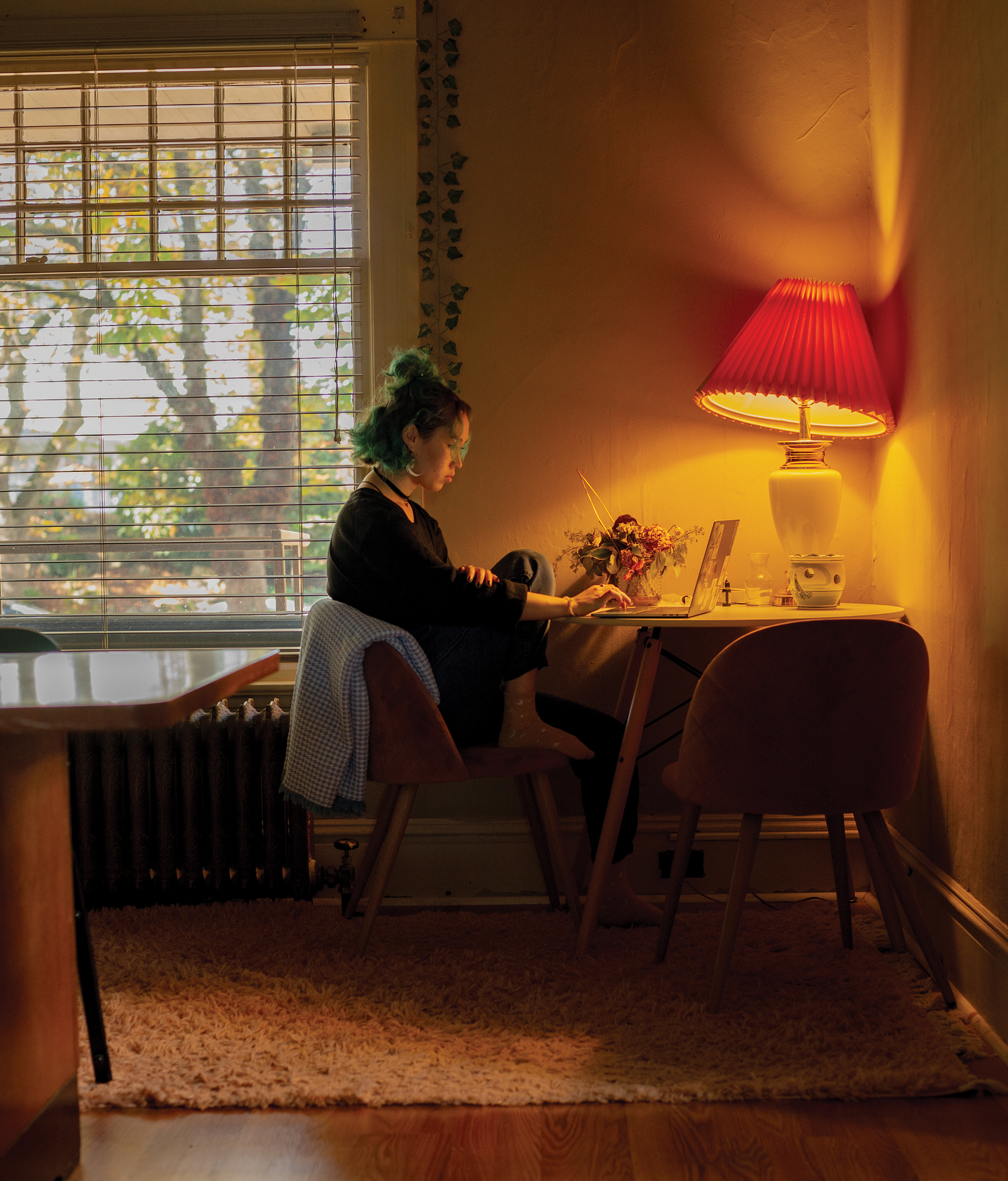 A student sits at home in a corner desk near a window using a laptop