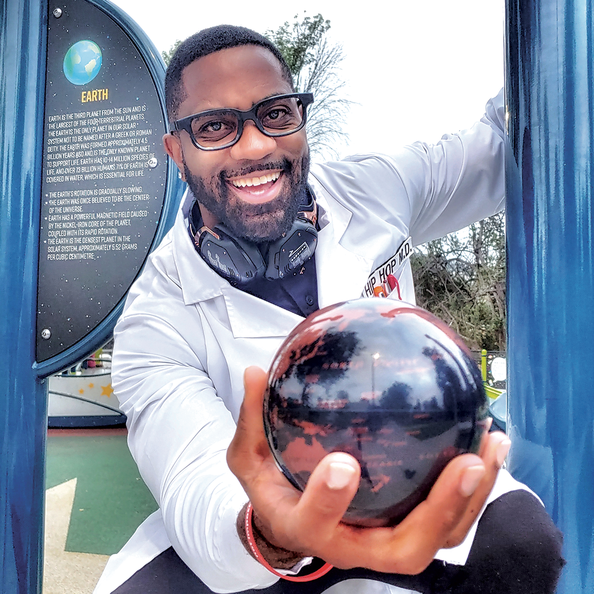 Maynard Okereke wearing a white coat and glasses, smiling, holding a small globe.