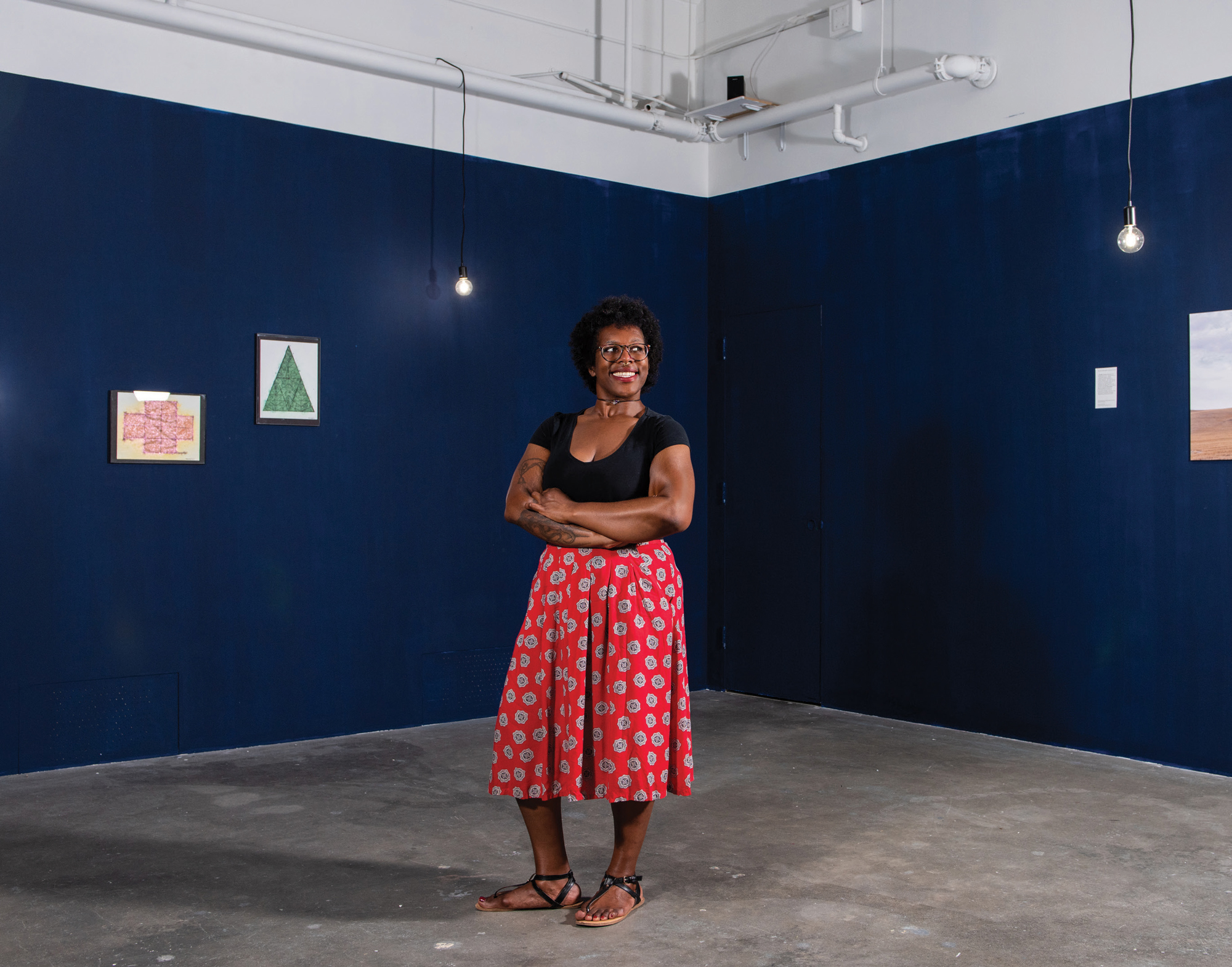 A smiling woman, wearing a black shirt and red patterned skirt, crosses her arms and looks off into the distance in an art gallery.
