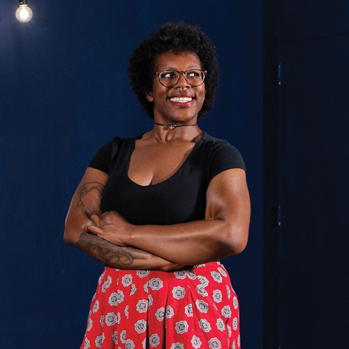 A smiling woman, wearing a black shirt and red patterned skirt, crosses her arms and looks off into the distance.