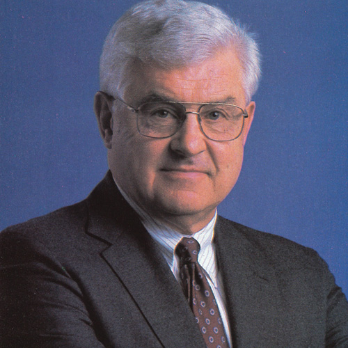 Headshot of Tracy King, a white-haired man in a grey suit with his arms crossed