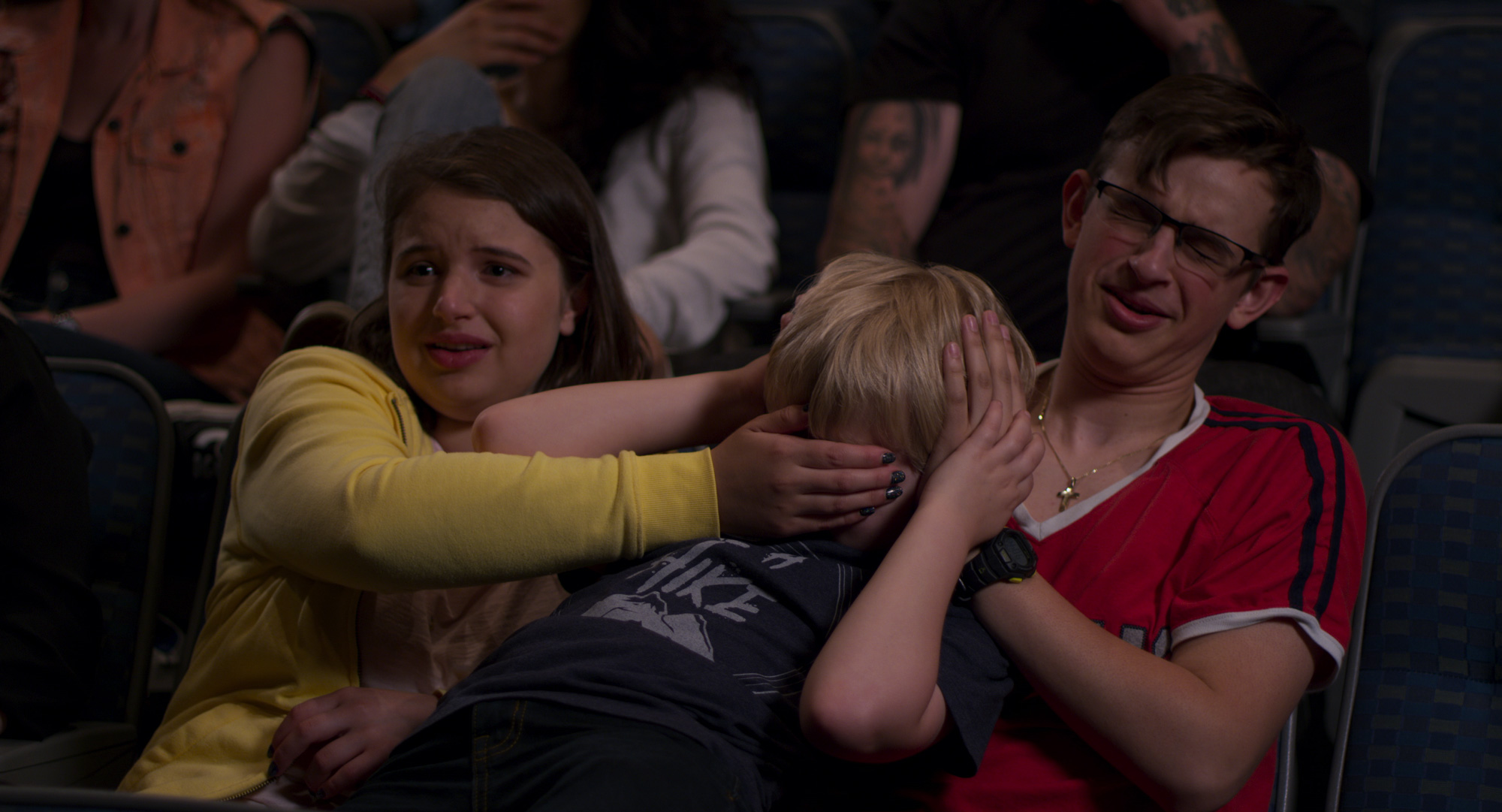 Two older children in theater seats cover the eyes and ears of a younger child.