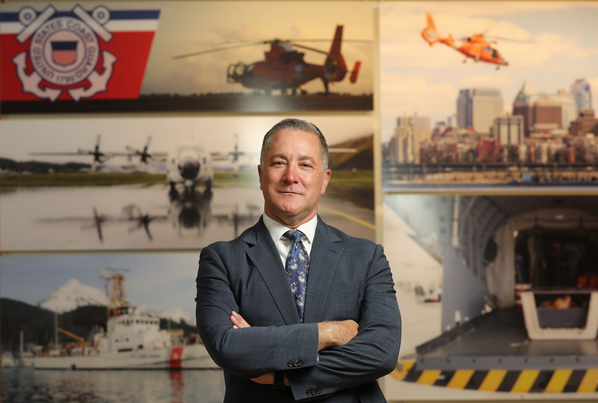 Michael Kilmer in a grey suit crossing his arms in front of a military mural
