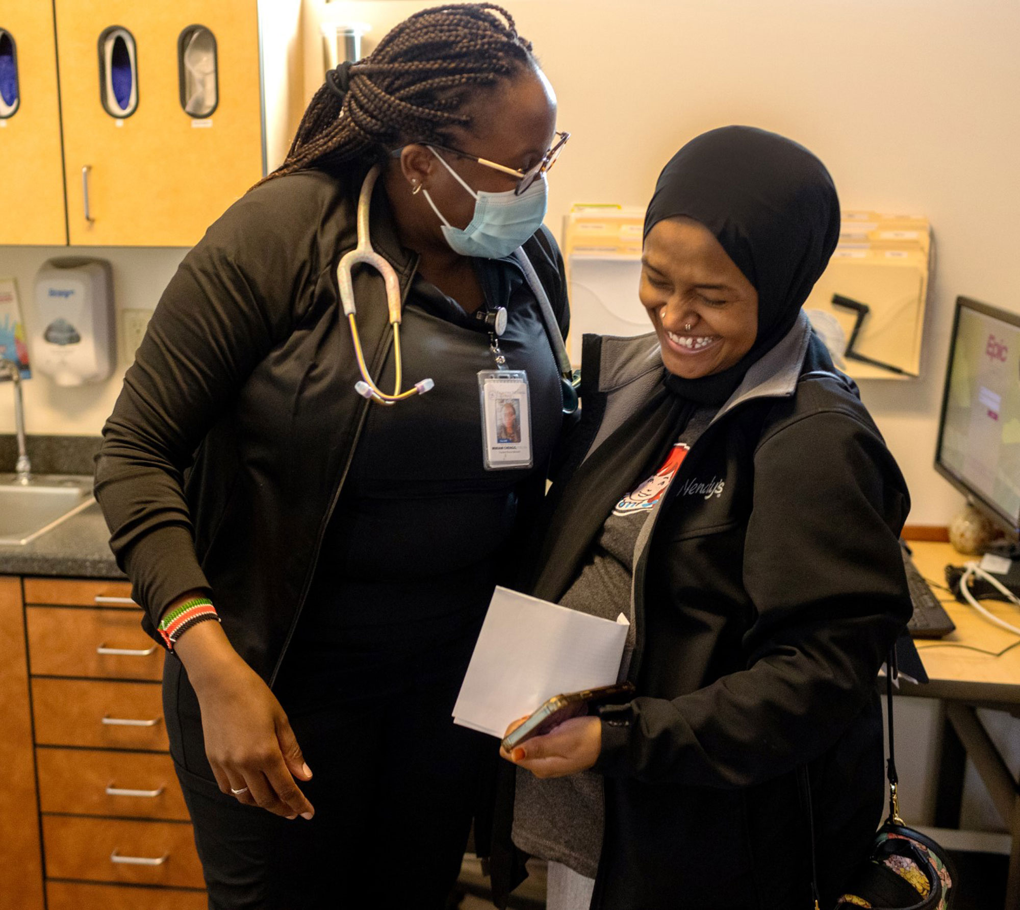 A woman wearing a surgical mask and stethoscope smiles at a laughing patient.