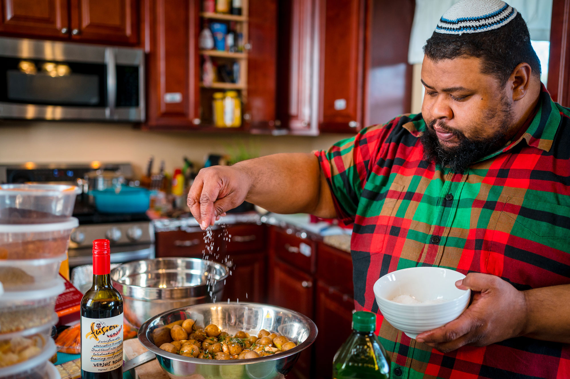 A Black man wearing a yarmulke sprinkles salt on potatoes in the kitchen.