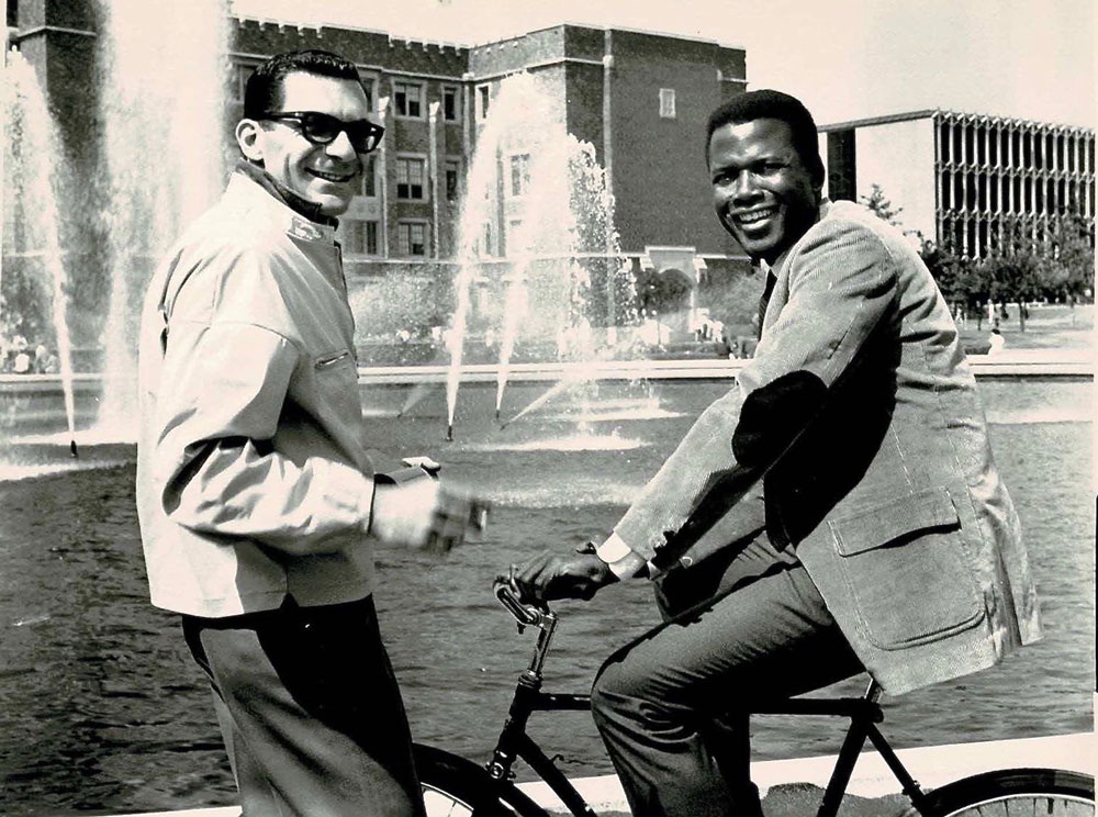 One man standing with sunglasses on, another sits on a bicycle, both smiling in front of a large fountain on the UW campus.