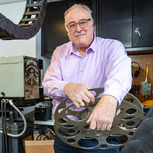A man smiles in the projection booth of a theater, holding a film reel.