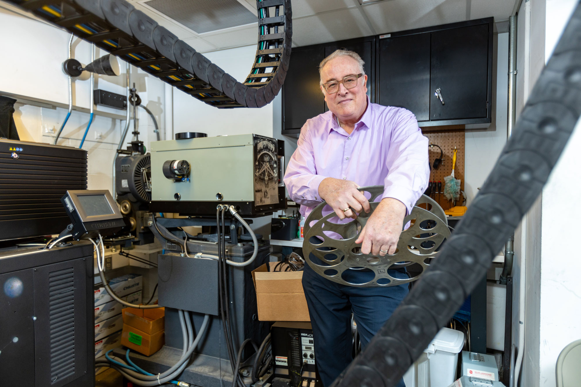 A man smiles in the projection booth of a theater, holding a film reel.