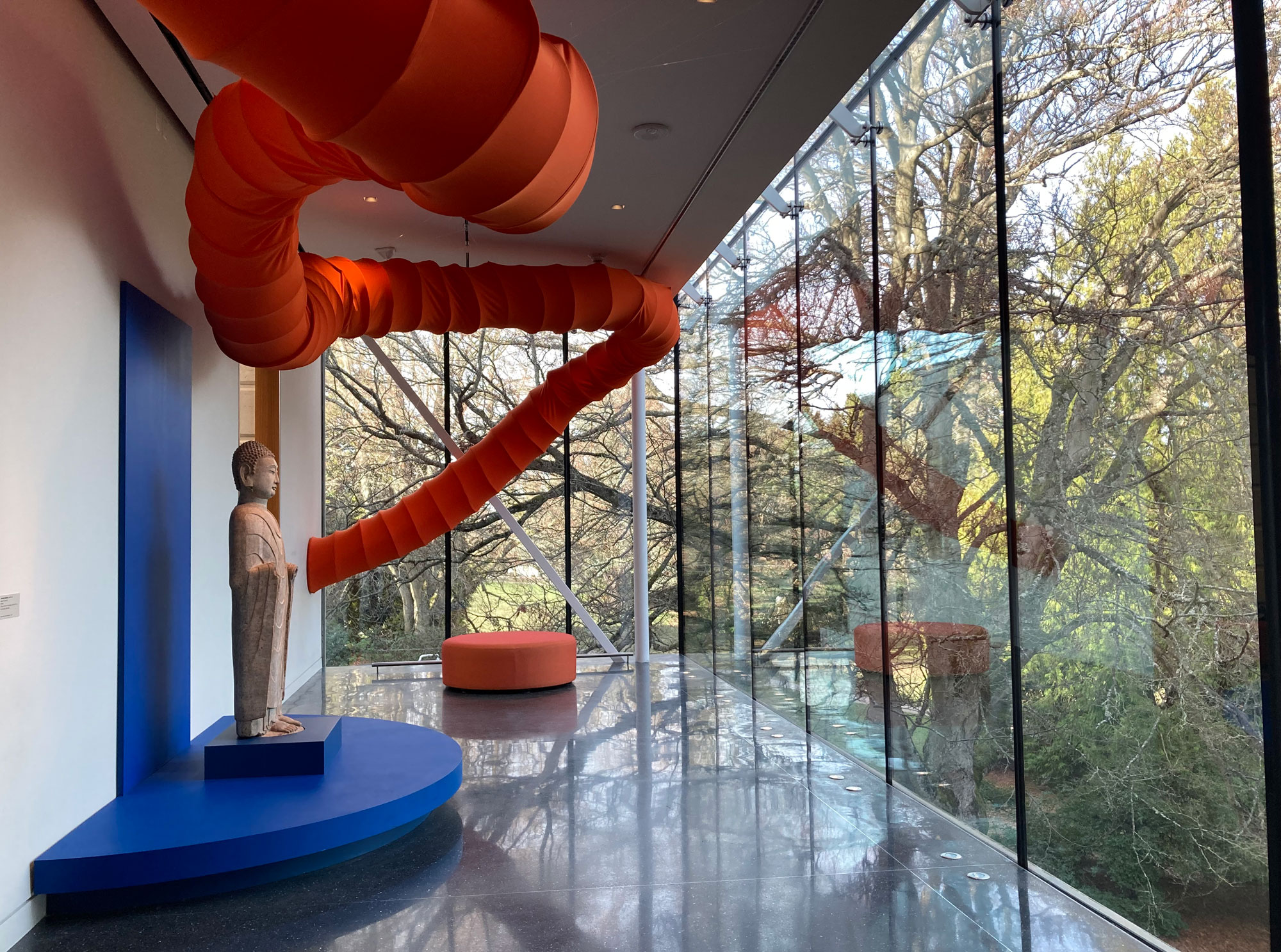 Inside an art museum, a statue of a buddha faces a wall of windows, and an orange tube winds its way down the hallway.