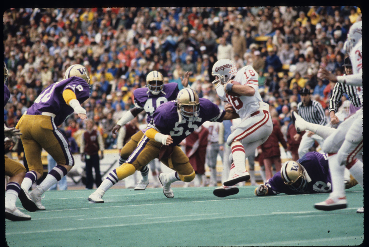 A vintage film photograph shows a group of football players in action, including Bruce Harrell in a UW football uniform shoving a man in a Washington State uniform.