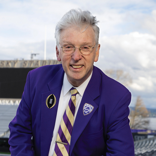 An older man wearing a purple suit and a striped tie smiles in Husky Stadium