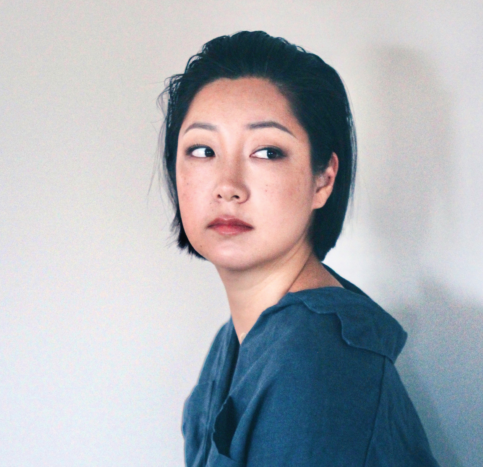 A young Korean woman with her dark hair slicked back stares over her shoulder with an inquisitive expression