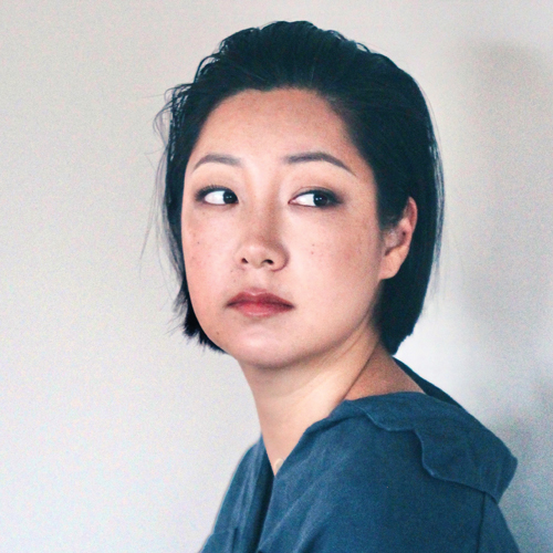 A young Korean woman with her dark hair slicked back stares over her shoulder with an inquisitive expression