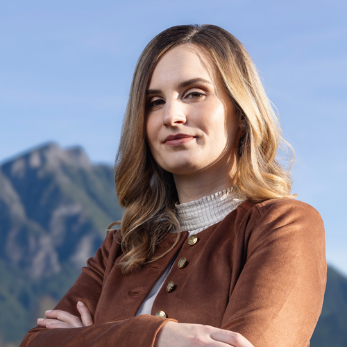 A woman with honey blonde hair, wearing a brown suede jacket, crosses her arms in a large field with mountains in the background