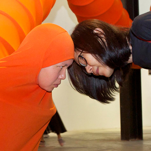 A museum visitor brings her fact close to Anida Ali, who is wearing a giant orange tube that winds around the room