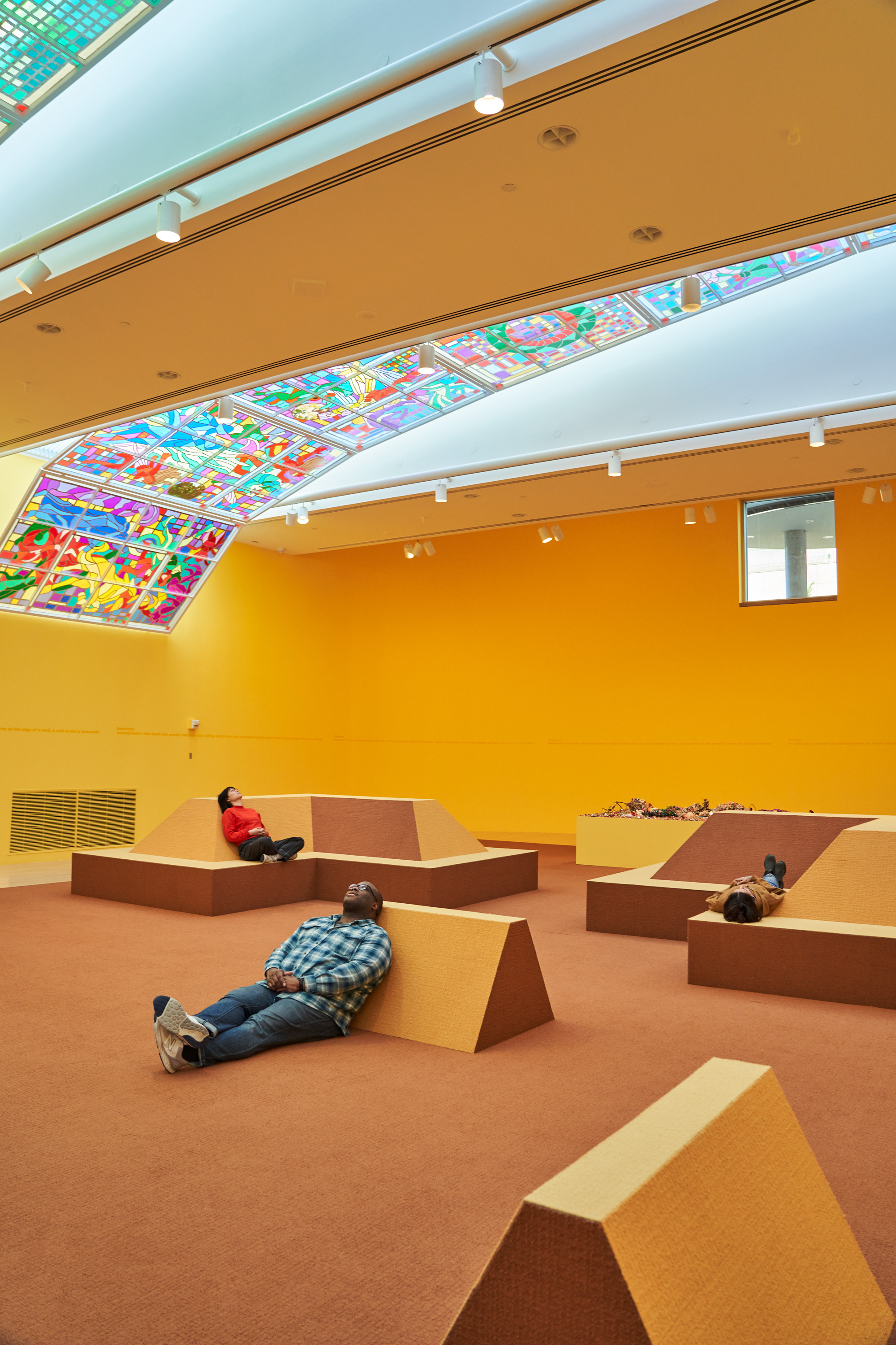 People relaxing in a bright yellow museum room. They're staring at the ceiling, which has a large panel of acetate and tape made to look like stained glass.