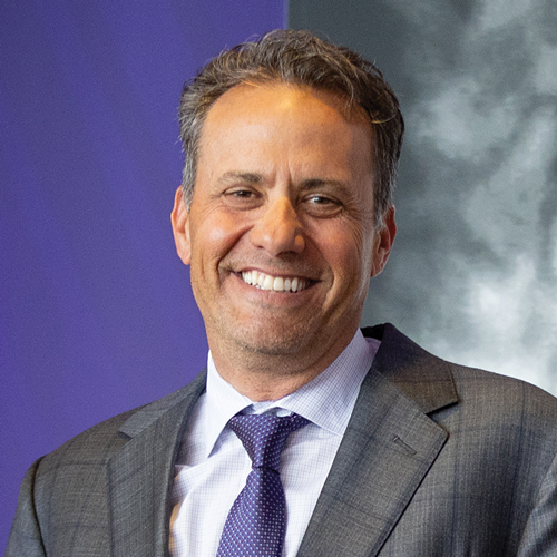 A man smiling and wearing a purple and white polka dot tie and gray suit