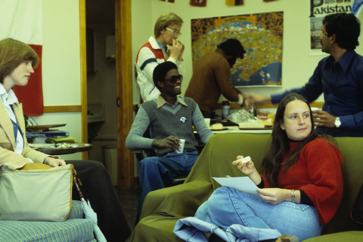 Six students - two white women, one white man and three men of color - lounge and eat snacks in the Hub on couches and chairs.