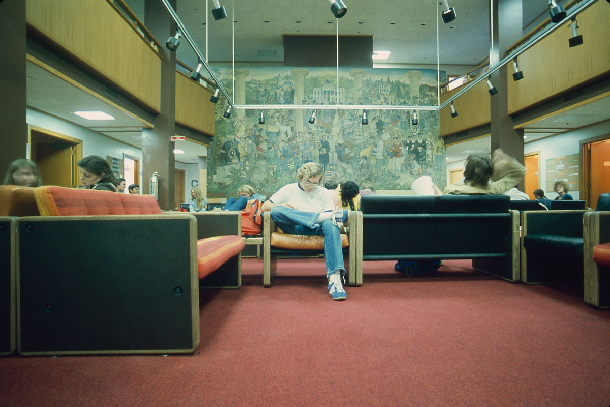 A blond-haired man wearing a t-shirt and jeans studies a book in the lobby of the Hub in the 1980s.