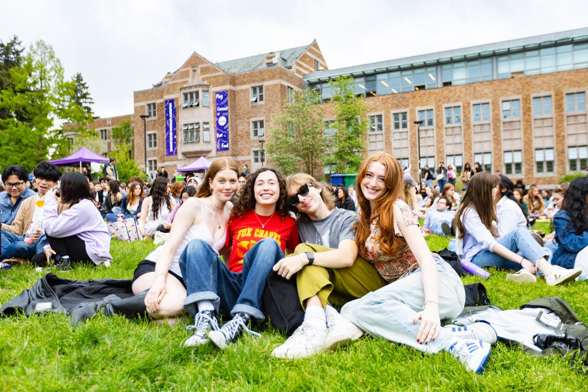Four young students sit in a crowd waiting for a concert on the Hub lawn. They are smiling and posing.