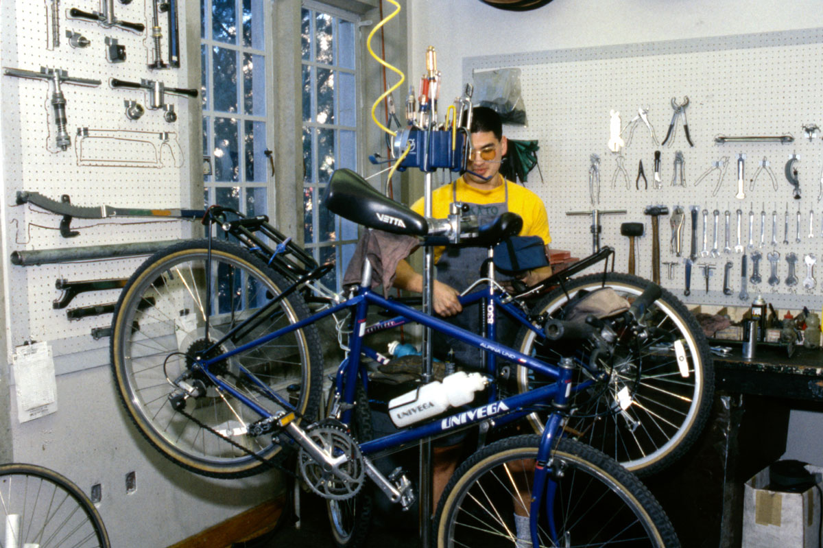 A man in a yellow shirt and denim apron works on a bicycle in the ASUW Bike Shop.