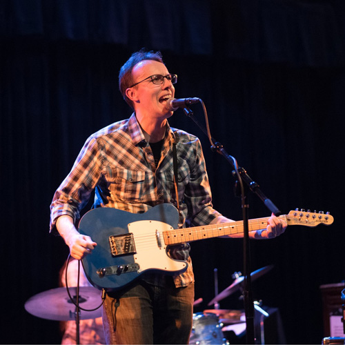 A man in a plaid shirt sings and performs on an electric guitar onstage