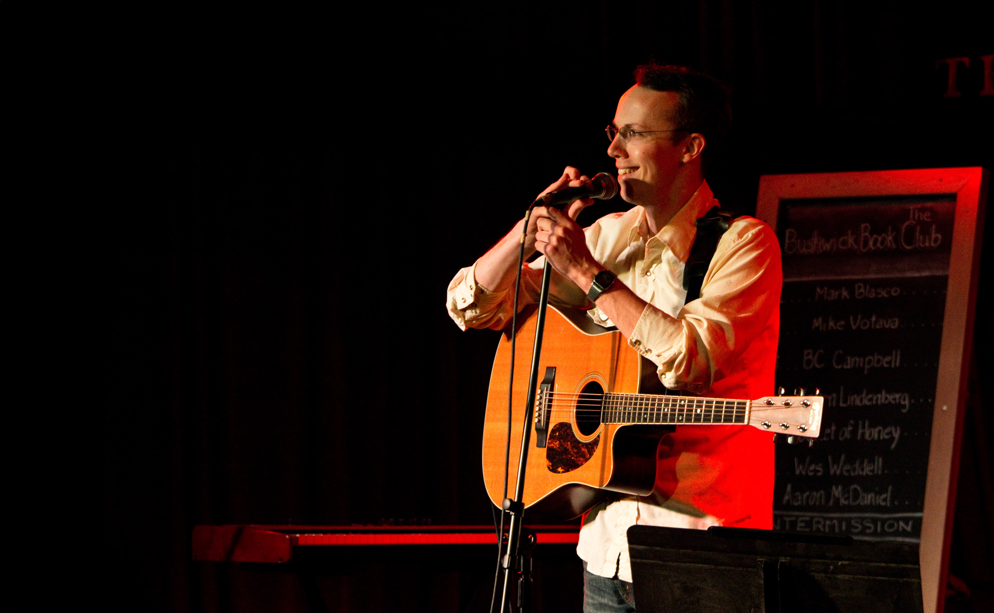 A man wearing a button-up shirt holds an acoustic guitar and speaks into a microphone onstage.
