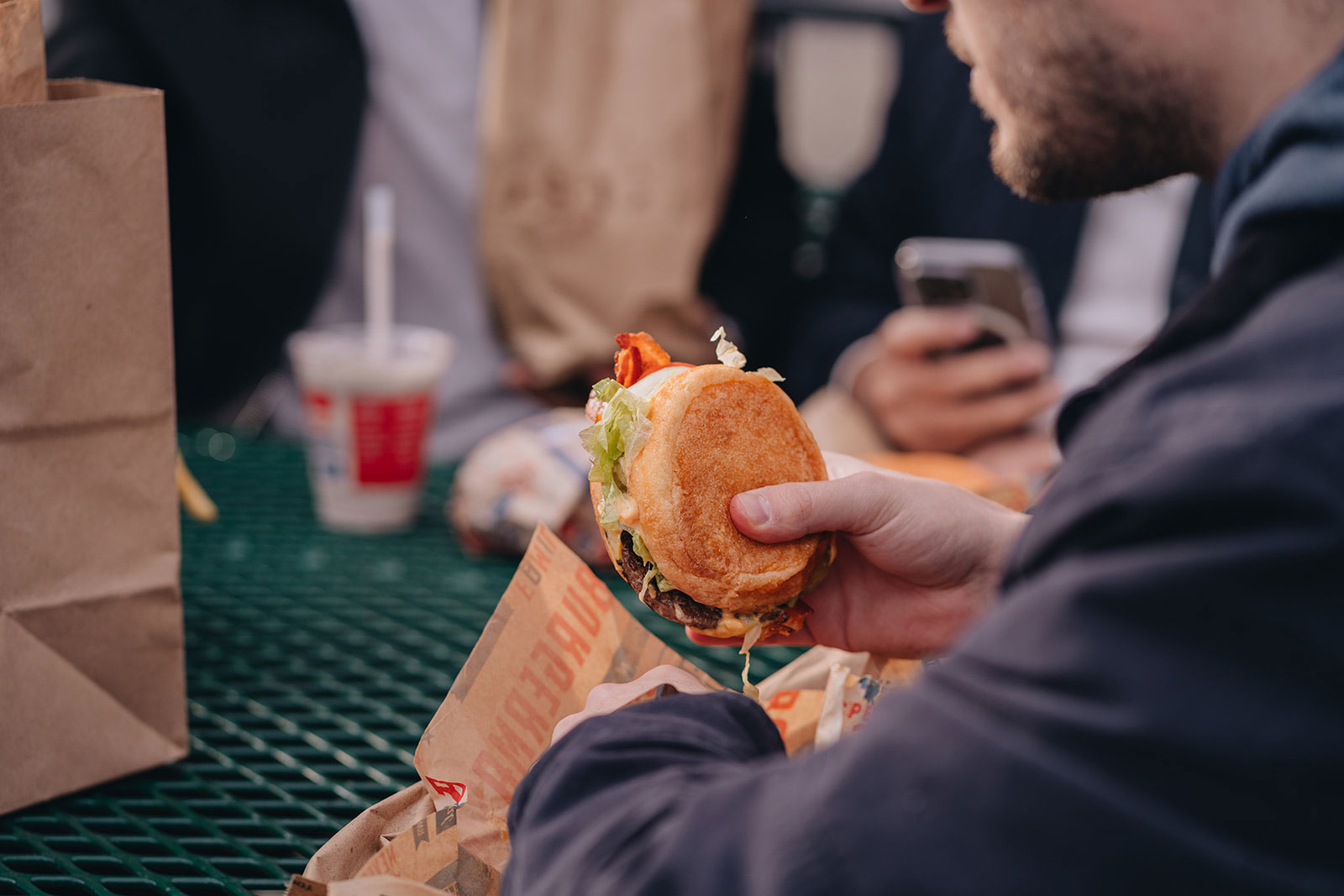 A man holds a burger at an outdoor table surrounded by other people also eating.