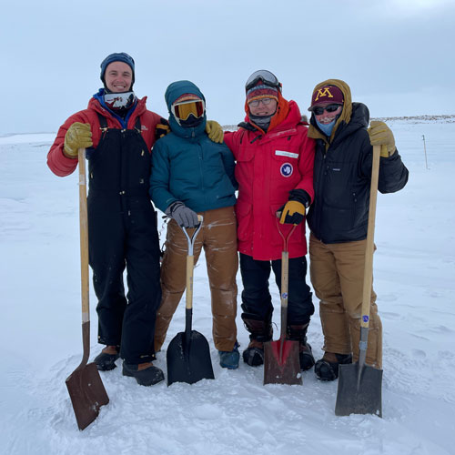 Four researchers pose wearing heavy winter gear and holding shovels in an icy landscape