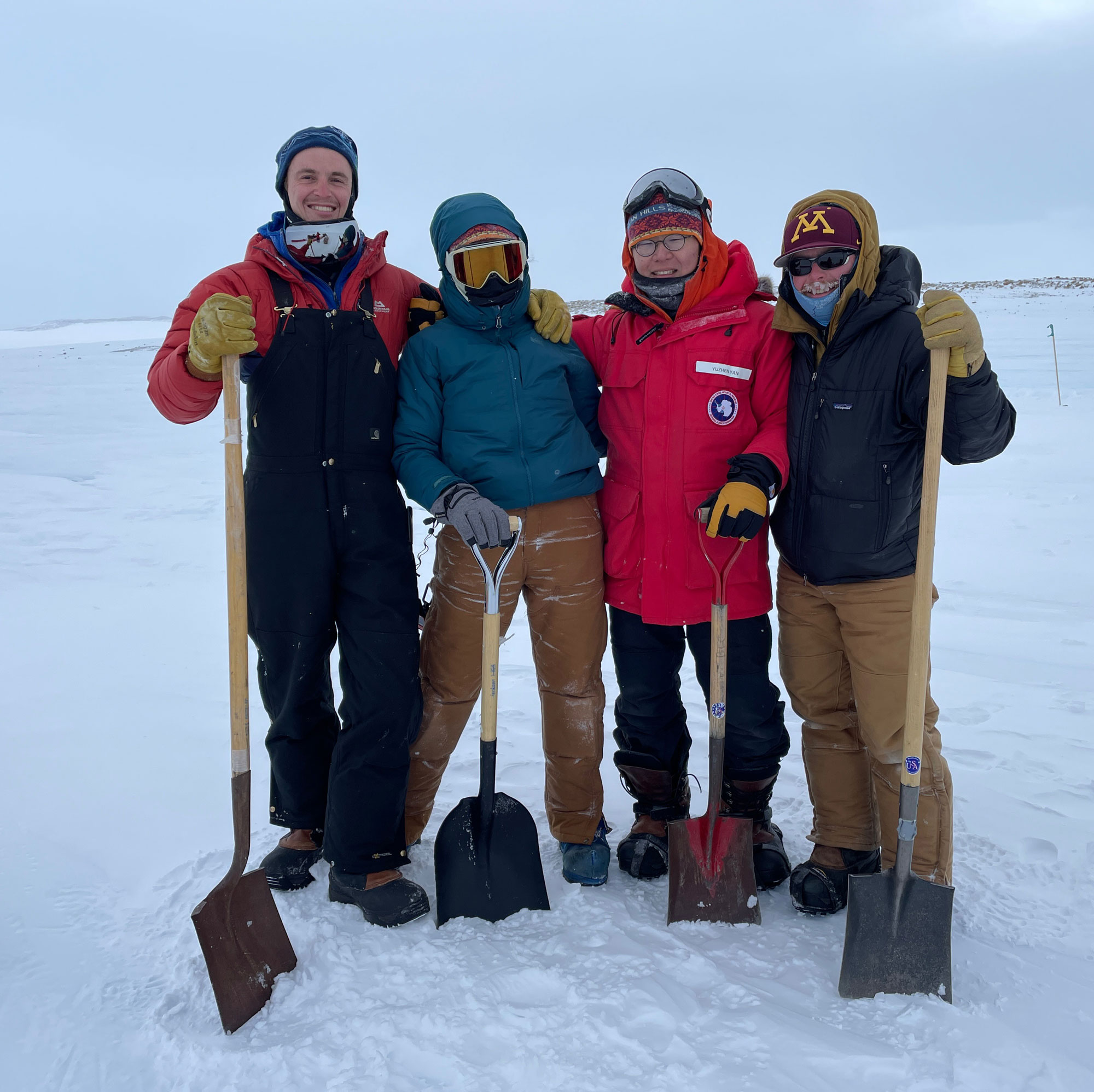 Four researchers pose wearing heavy winter gear and holding shovels in an icy landscape