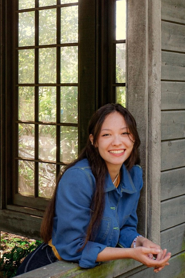 A long-haired young woman in a denim shirt smiles, leaning out of a barn window opening