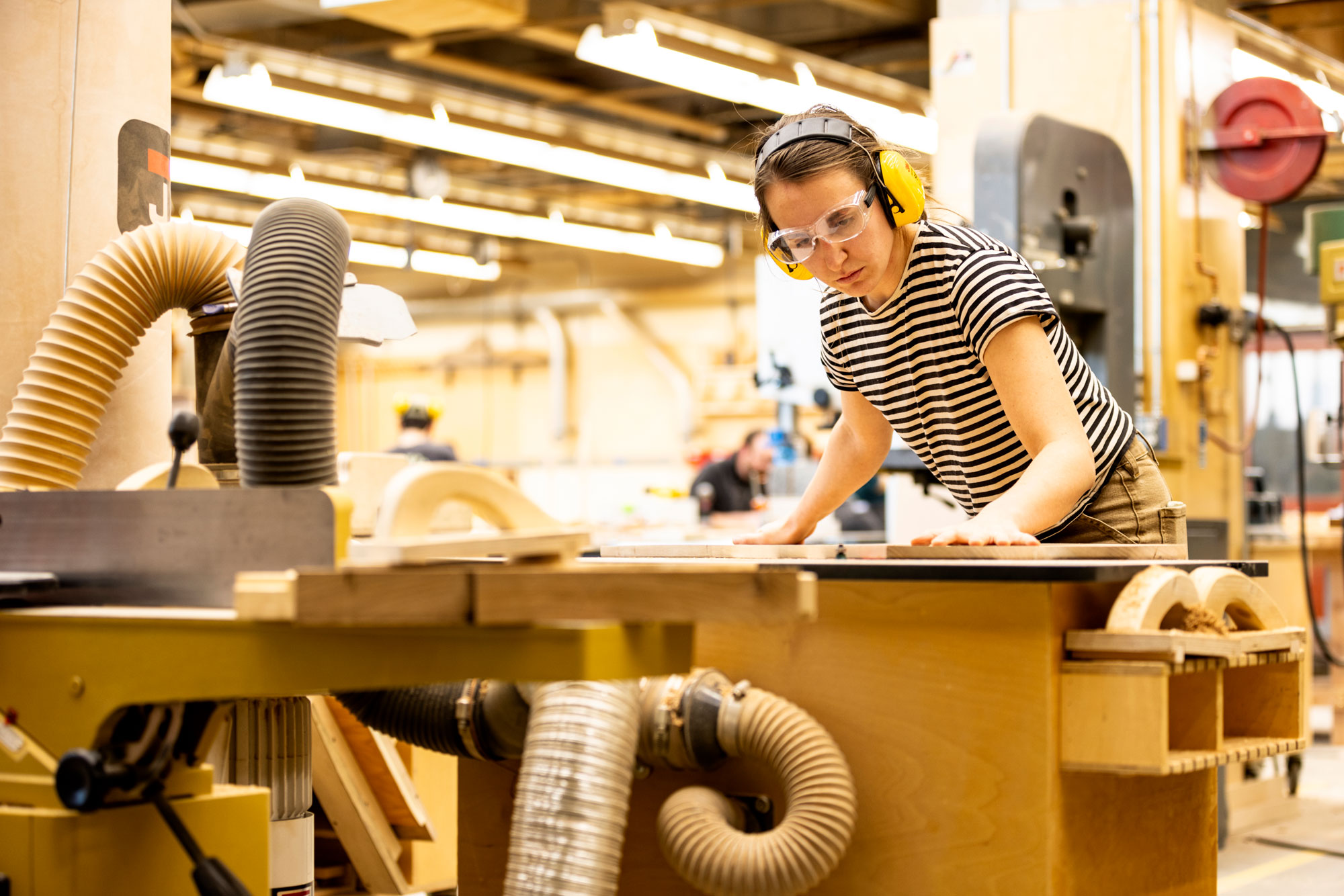 A woman in a striped shirt and protective gear steadies a piece of wood in a workshop