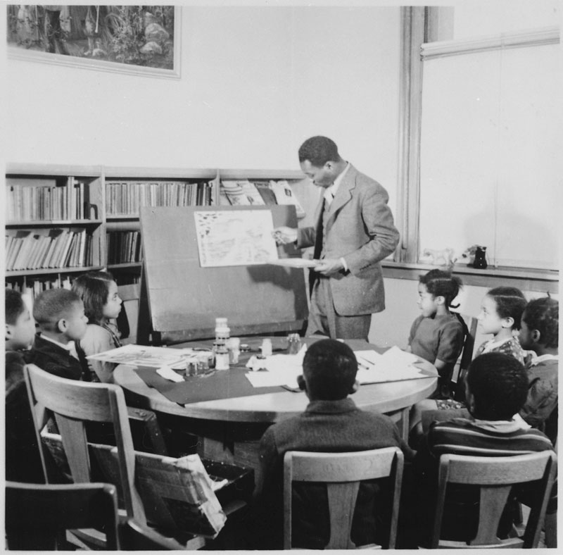 An old black and white photo of a man in a suit teaching a small classroom of children