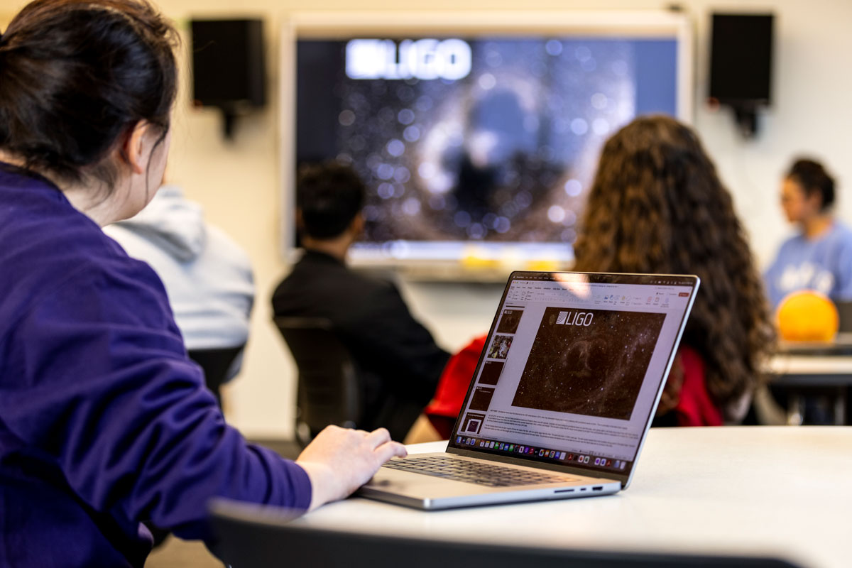 A woman in a purple sweatshirt takes notes on her computer while an astronomy professor lectures.