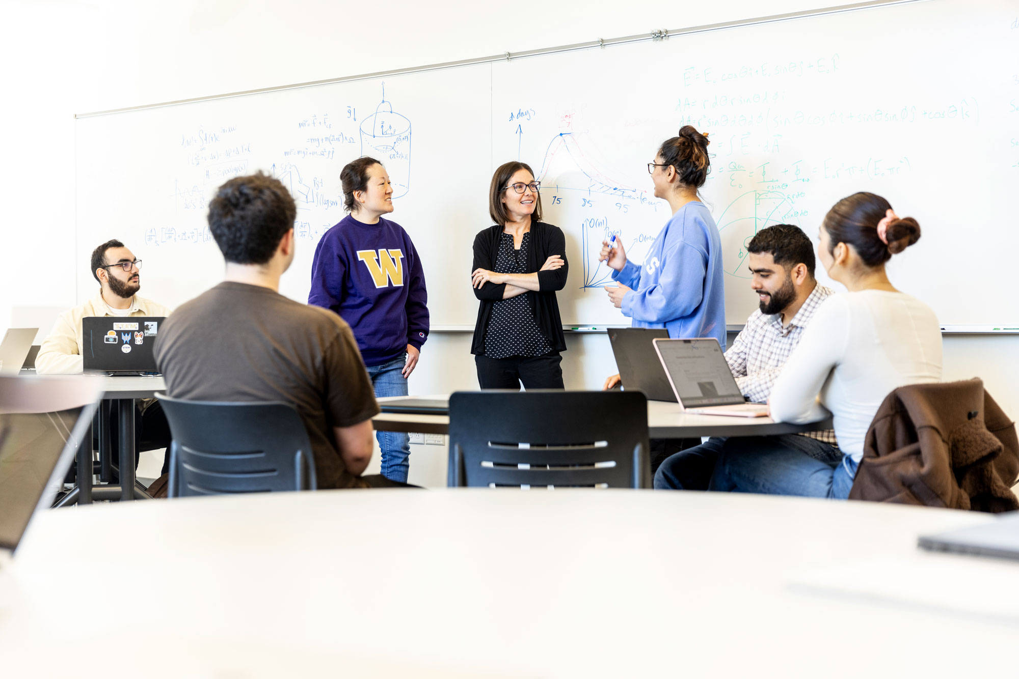 Joey Kay, a professor wearing a blouse and cardigan with black pants, stands in front of a whiteboard chatting with several students